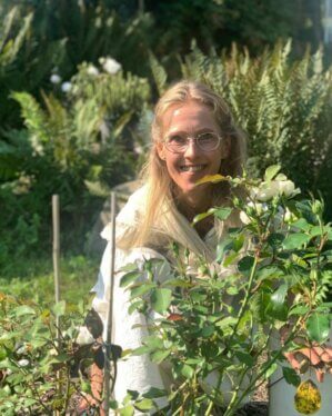 Portrait einer Frau mit blonden Haaren und Brille. Sie steht hinter Pflanzen im Garten